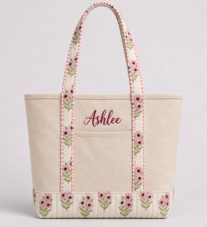 May include: A beige canvas tote bag featuring the name Ashlee embroidered in dark pink script. The bag has two handles and a bottom panel decorated with a repeating pattern of pink flowers and green leaves, accented by a pink and white gingham trim.