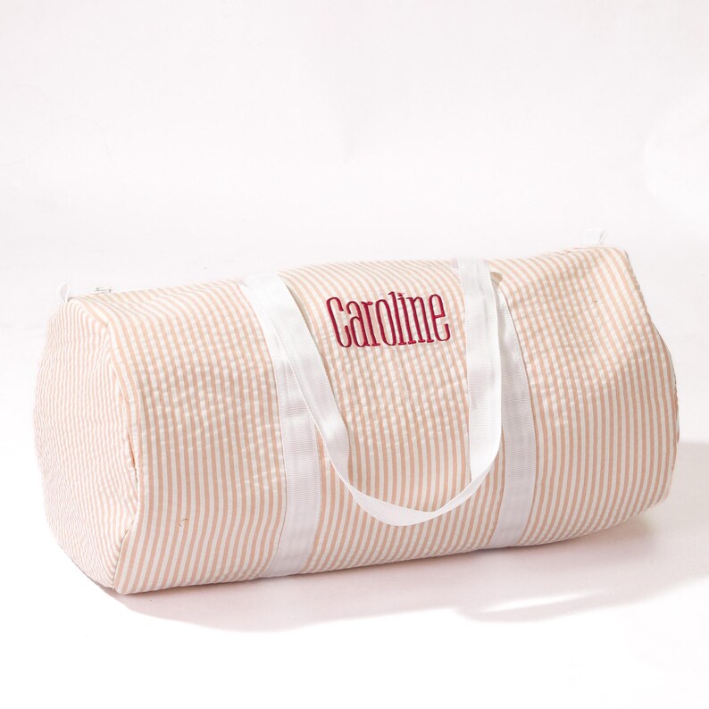 Puede incluir: Una bolsa de viaje de lona a rayas rosa y blancas con asas blancas y el nombre "Caroline" bordado en rojo.