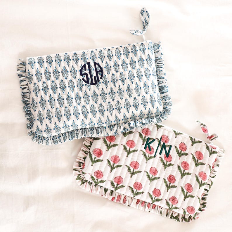 May include: Two quilted cosmetic bags. One is white with a blue floral pattern and the initials "SLA" embroidered in navy. The other is white with a pink floral pattern and the initials "K/N" embroidered in green. Both have ruffled edges.