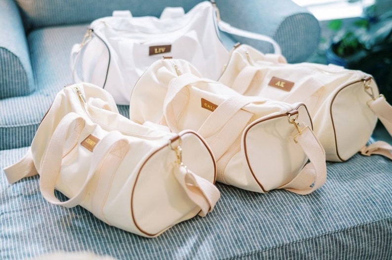 K&ouml;nnte beinhalten: Vier wei&szlig;e Canvas-Reisetaschen mit braunen Lederbes&auml;tzen und goldenen Rei&szlig;verschluss-Z&uuml;gen. Jede Tasche hat einen Namensmonogramm auf der Vorderseite: Liv, Cal, AK und Carey.