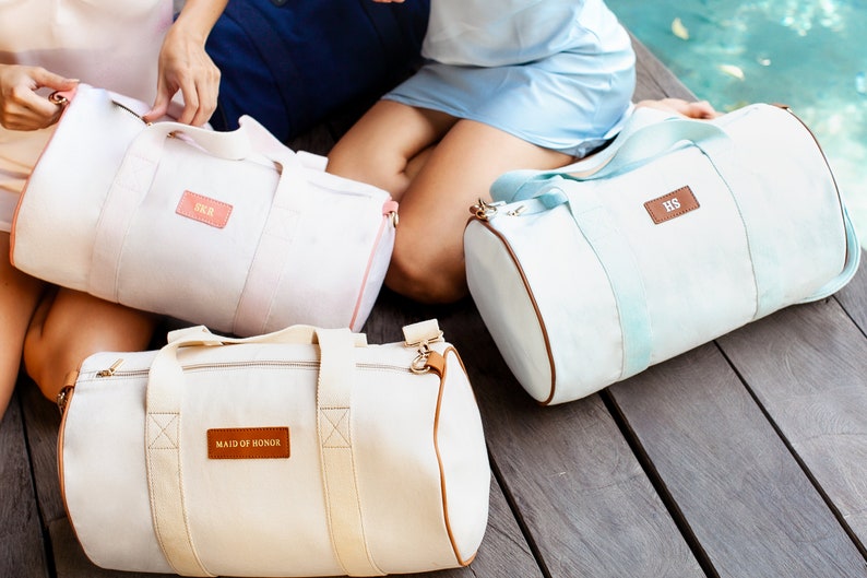 K&ouml;nnte beinhalten: Drei personalisierte Canvas-Reisetaschen in Wei&szlig;, Hellrosa und Hellblau. Die Taschen haben braune Lederakzente und personalisierte Etiketten mit dem Text "Maid of Honor", "SKR" und "HS".