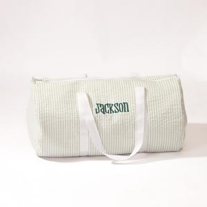 Puede incluir: Una bolsa de viaje de lona a rayas blancas y verdes con asas blancas y el nombre "Jackson" bordado en verde.