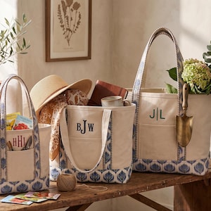 May include: Three off-white canvas tote bags with blue floral patterned trim at the base and handles. Each bag features a front pocket and embroidered monogram initials in dark blue or red thread, containing gardening tools and accessories.