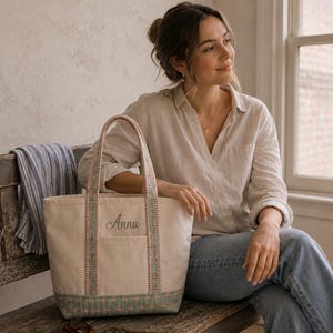 May include: A cream-colored canvas tote bag featuring the name Anna embroidered in dark gray script on the front pocket. The bag has two handles and a base panel decorated with a green and pink quilted pattern, with matching trim along the straps.