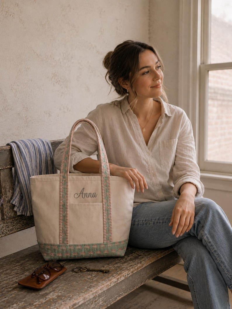 May include: A cream-colored canvas tote bag featuring the name Anna embroidered in dark gray script on the front pocket. The bag has two handles and a base panel decorated with a green and pink quilted pattern, with matching trim along the straps.