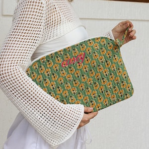 Puede incluir: Clutch acolchado verde con un estampado floral en amarillo y naranja. El clutch tiene un nombre bordado en rosa y un asa peque&ntilde;a. El fondo es una pared blanca.