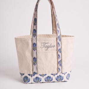May include: A cream-colored canvas tote bag featuring a blue and green floral block print pattern on the base and handles. The bag includes two vertical straps and the name Taylor embroidered in a cursive script on the front pocket area.