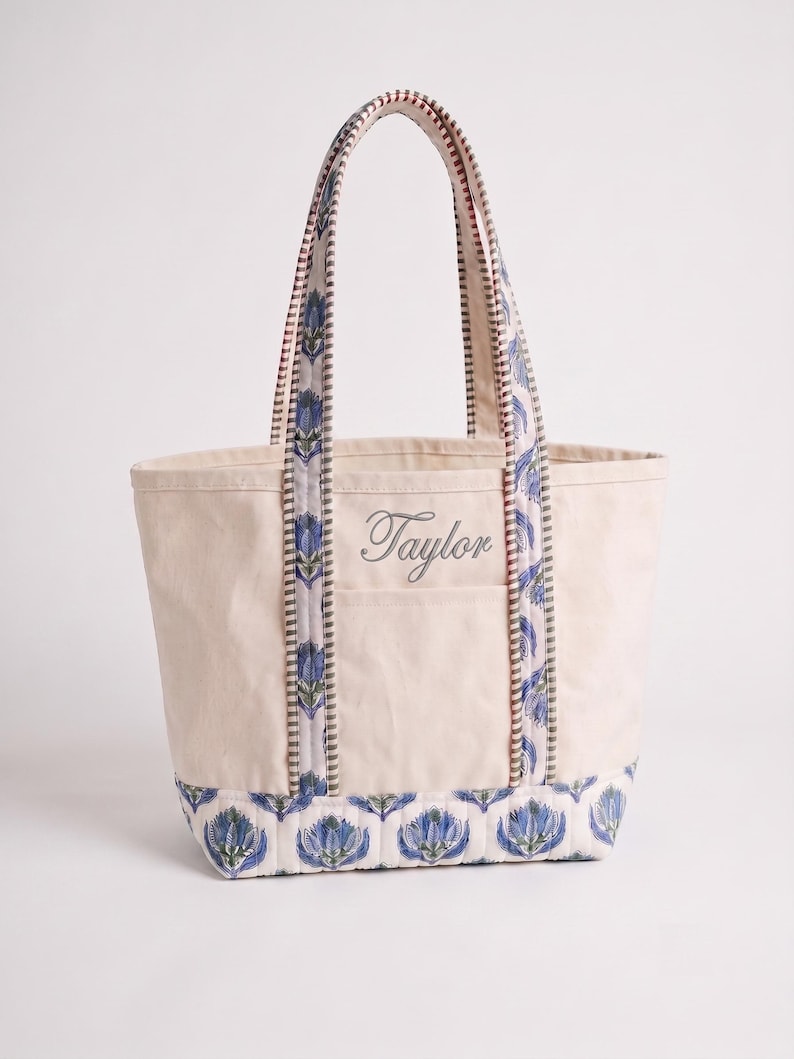 May include: A cream-colored canvas tote bag featuring a blue and green floral block print pattern on the base and handles. The bag includes two vertical straps and the name Taylor embroidered in a cursive script on the front pocket area.