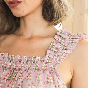 May include: A light purple sleeveless top featuring a repeating floral pattern of small pink flowers and green stems. The garment has a square neckline with gathered ruffles along the shoulder straps and a small embroidered monogram in gold thread.