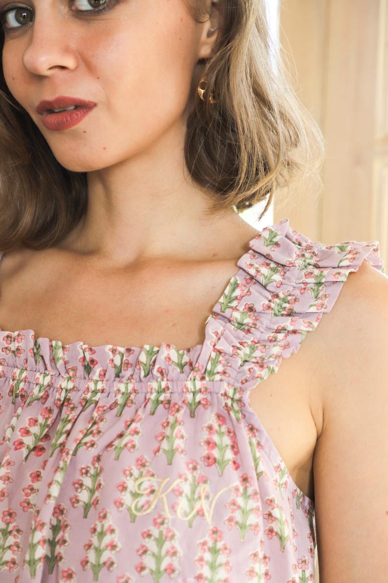 May include: A light purple sleeveless top featuring a repeating floral pattern of small pink flowers and green stems. The garment has a square neckline with gathered ruffles along the shoulder straps and a small embroidered monogram in gold thread.