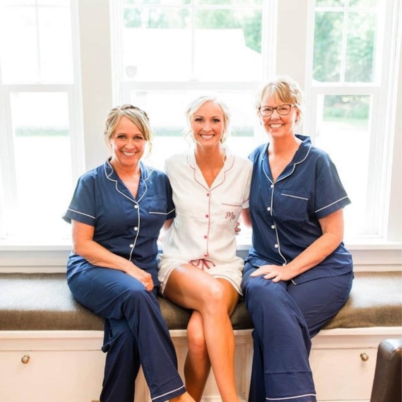 May include: Three women wearing matching pajamas, two in navy blue and one in white, with white piping and a red button detail. The women are sitting on a window seat, smiling at the camera.