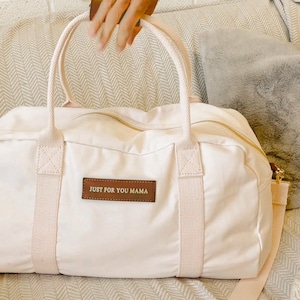 May include: A white canvas duffel bag with brown leather accents and a gold zipper. The bag has two top handles and a long shoulder strap. The bag has a brown leather patch with the text "JUST FOR YOU MAMA" in gold lettering.
