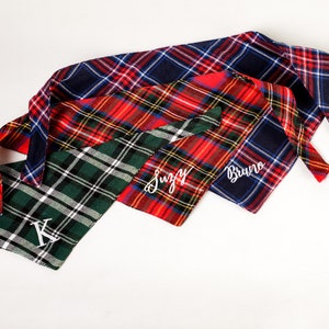 Pu&ograve; includere: Tre bandane per cani a quadri con nomi ricamati in bianco. Le bandane sono rosso e nero, verde e bianco e blu e rosso. I nomi sono "Bruno", "Suzy" e "K".