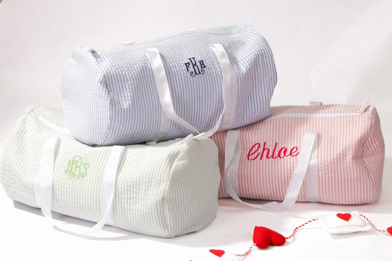 Puede incluir: Tres bolsas de lona rayadas con correas blancas. Las bolsas est&aacute;n personalizadas con los nombres "Chloe", "F.B." y un monograma.