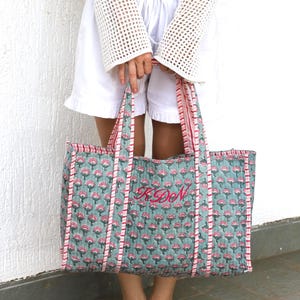 May include: A large tote bag with a floral pattern in teal and pink. The bag has pink striped trim and handles. The initials "KDN" are embroidered in pink on the front. The bag is held by a person.
