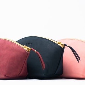 May include: Three leather zippered pouches in different colors: burgundy, black, and pink. Each pouch has a gold zipper and a small leather pull tab. The pouches are lined up in a row, with the burgundy pouch on the left, the black pouch in the middle, and the pink pouch on the right.