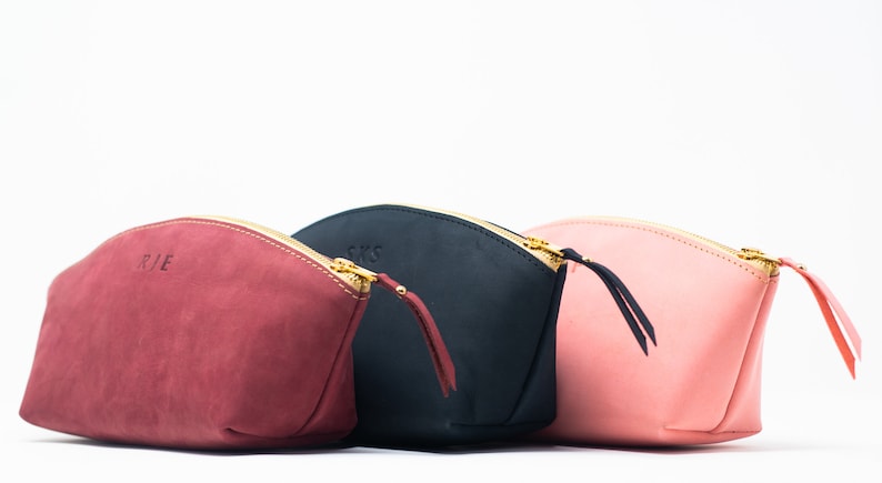 May include: Three leather zippered pouches in different colors: burgundy, black, and pink. Each pouch has a gold zipper and a small leather pull tab. The pouches are lined up in a row, with the burgundy pouch on the left, the black pouch in the middle, and the pink pouch on the right.