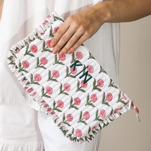 May include: White quilted clutch with a floral design in pink and green. The clutch features a ruffled edge and is monogrammed with the initials "KN" in green. The clutch is held by a person wearing a white outfit.
