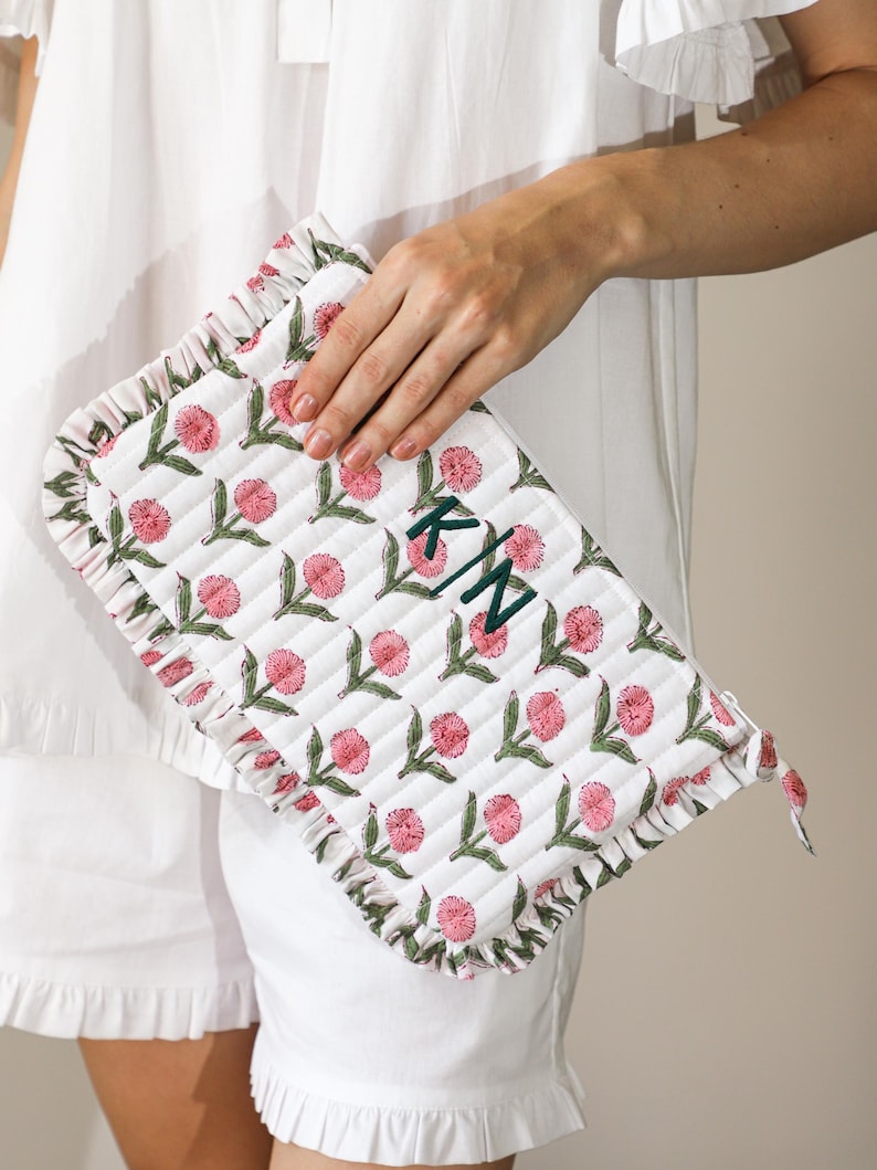 May include: White quilted clutch with a floral design in pink and green. The clutch features a ruffled edge and is monogrammed with the initials "KN" in green. The clutch is held by a person wearing a white outfit.