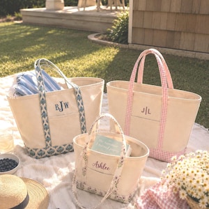 May include: Three off-white canvas tote bags with open tops and front pockets. One bag features blue patterned trim and the monogram BJW. Another has pink gingham trim and the monogram J|L. The smallest bag has floral trim and the name Ashlee.