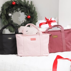 May include: Three tote bags in different colors: black, pink, and burgundy. Each bag has a simple design with two handles and a small label on the front.
