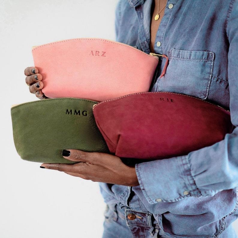 May include: Three personalized leather cosmetic bags in pink, green, and burgundy. The bags are stacked on top of each other and held by a person. The bags have the initials "ARZ", "MMG", and "RJE" embossed on them.
