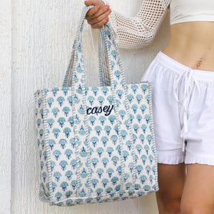 May include: A blue and white floral patterned tote bag with the name "casey" embroidered in black. The bag has matching blue and tan striped handles. The bag is held in front of a white textured wall.