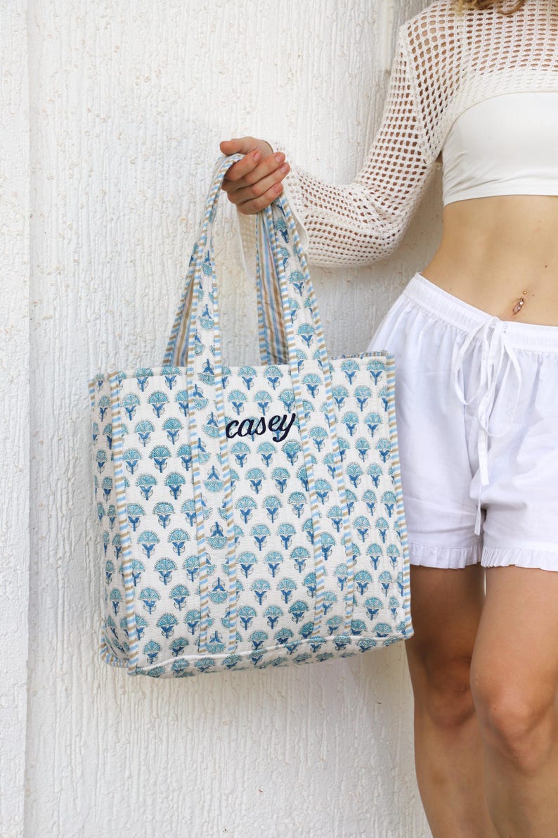 May include: A blue and white floral patterned tote bag with the name "casey" embroidered in black. The bag has matching blue and tan striped handles. The bag is held in front of a white textured wall.
