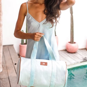 K&ouml;nnte beinhalten: Eine hellblaue Canvas-Sporttasche mit braunen Lederdetails und einem personalisierten "HS"-Monogramm. Die Tasche wird von einer Person getragen, die ein hellblaues Schlupfkleid tr&auml;gt.