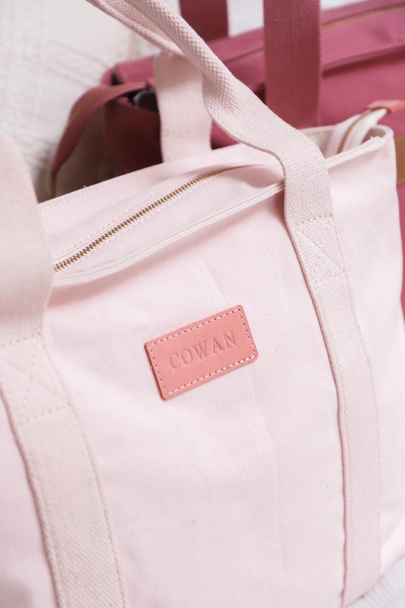 May include: A pale pink canvas tote bag with a pink leather patch that says "COWAN" on it. The bag has a zipper closure and two long straps.