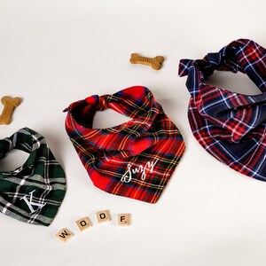 Pu&ograve; includere: Tre bandane per cani personalizzate in tartan rosso, verde e blu con nomi ricamati in bianco "Suzy", "Bruno" e "K". Le bandane sono disposte su una superficie bianca con dolcetti a forma di osso per cani e la parola "WOOF" scritta con tessere di lettere di legno.