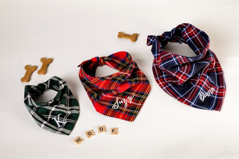 Pu&ograve; includere: Tre bandane per cani personalizzate in tartan rosso, verde e blu con nomi ricamati in bianco "Suzy", "Bruno" e "K". Le bandane sono disposte su una superficie bianca con dolcetti a forma di osso per cani e la parola "WOOF" scritta con tessere di lettere di legno.