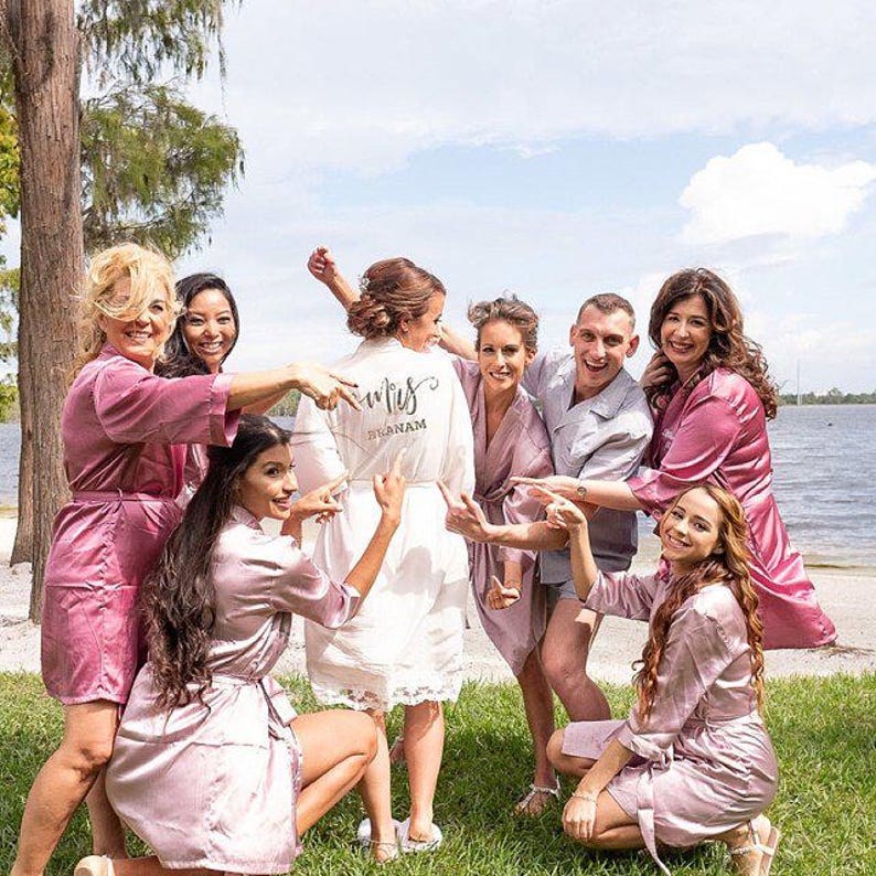 May include: A group of people, some wearing pink satin robes, gather outdoors near a body of water. One person wears a white robe with the word "Mrs" and a name on the back. The scene suggests a wedding or bridal event.
