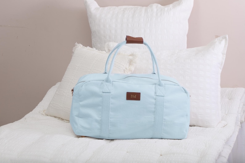 K&ouml;nnte beinhalten: Eine hellblaue Reisetasche aus Canvas mit braunen Ledergriffen und einem braunen Lederetikett mit den Initialen "BM".