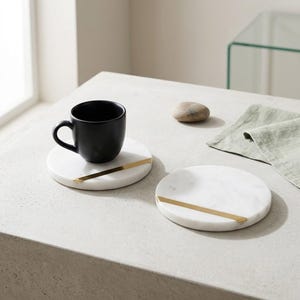 May include: A matte black ceramic mug sits on a circular white marble coaster featuring a thin, diagonal gold-tone metal inlay. Beside it, a second identical marble coaster with a gold-tone inlay rests on the surface, along with a smooth pebble.