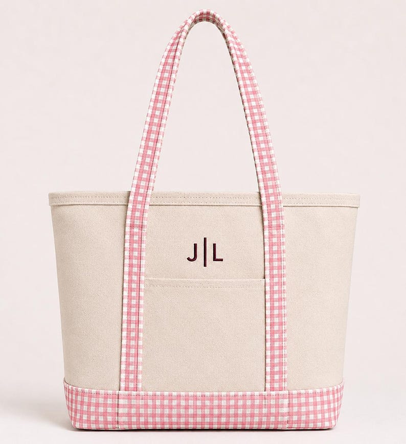 May include: A beige canvas tote bag featuring pink and white gingham patterned handles and a matching gingham base. The front of the bag includes an open pocket and is embroidered with the dark brown monogram letters J|L centered above the pocket.