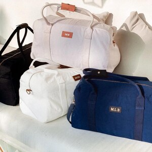 Puede incluir: Cuatro bolsas de viaje de lona con asas de cuero. Las bolsas son de color negro, blanco y azul marino. Las bolsas tienen nombres personalizados: "SAM", "WADE" y "M.L.B."