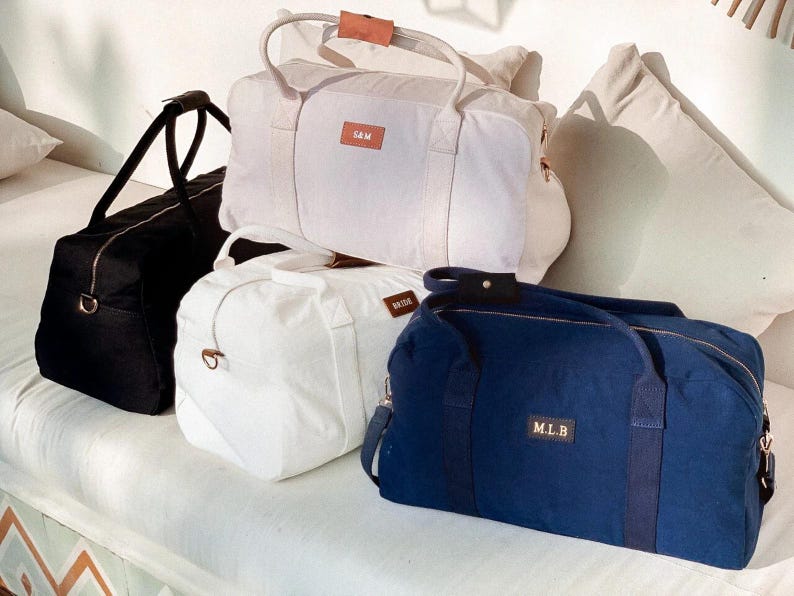 Puede incluir: Cuatro bolsas de viaje de lona con asas de cuero. Las bolsas son de color negro, blanco y azul marino. Las bolsas tienen nombres personalizados: "SAM", "WADE" y "M.L.B."