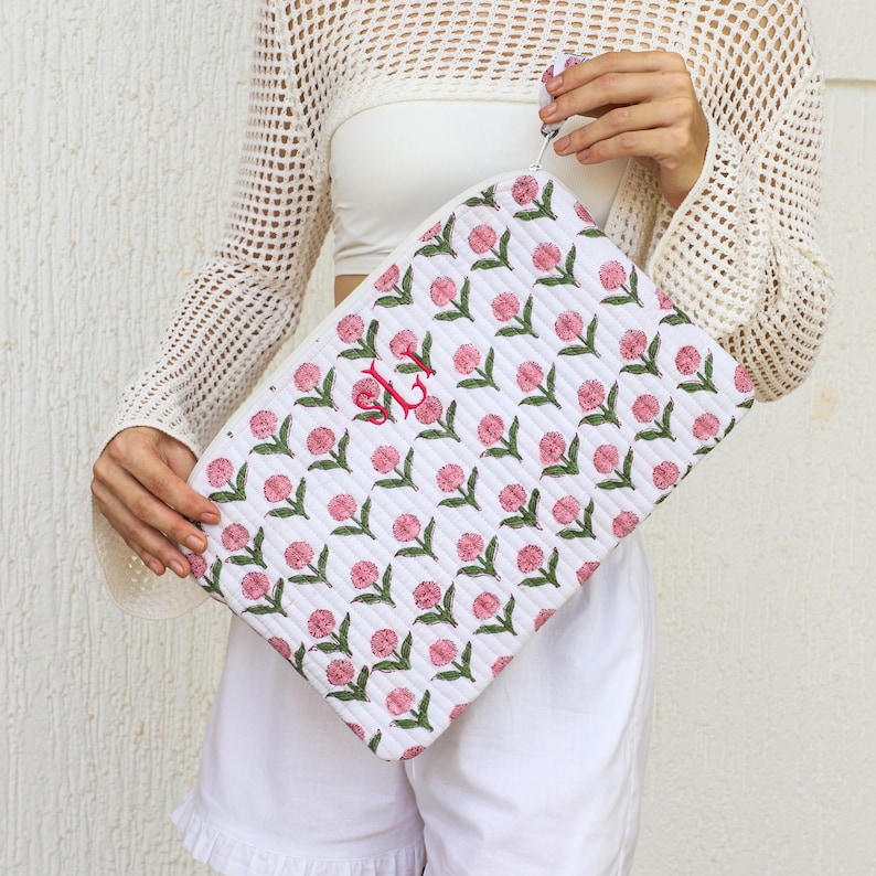 Puede incluir: Clutch acolchado blanco con un estampado floral rosa repetido y hojas verdes. El clutch presenta un monograma rojo y un asa peque&ntilde;a. La bolsa es sostenida por una persona que viste un atuendo blanco.