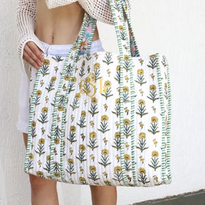 May include: White quilted tote bag with a floral pattern in yellow and green. The bag has green striped trim and handles with a floral design. Gold initials are embroidered on the front.