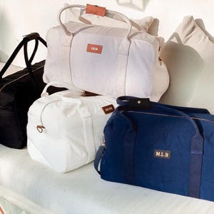 K&ouml;nnte beinhalten: Vier Reisetaschen aus Canvas mit Ledergriffen. Die Taschen sind in Schwarz, Wei&szlig; und Marineblau. Die Taschen haben personalisierte Namen: "SAM", "WADE" und "M.L.B."