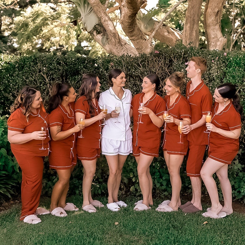 May include: A group of people wearing matching red pajamas with white trim and holding glasses of orange juice. They are standing in front of a green hedge.