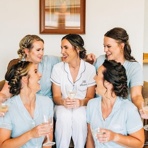 May include: A group of women in light blue and white pyjamas, laughing and holding glasses of champagne. One woman wears white pyjamas with black trim and the name "Mrs. Sheridan" embroidered on the shirt.