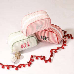 May include: Three striped fabric cosmetic bags with zipper closures. The bags are pink, red, and green with white stripes. Each bag has a monogram embroidered on the front. The bags are arranged on a white surface with a red garland.