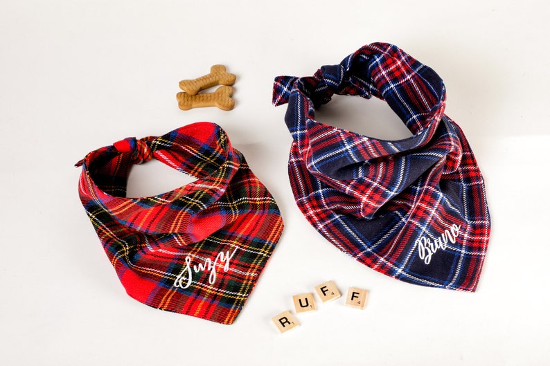 Pu&ograve; includere: Due bandane per cani a quadri con nomi personalizzati, "Suzy" e "Bruno", ricamate in bianco. Le bandane sono rosse e blu con un motivo a quadri bianchi. La parola "RUFF" &egrave; scritta con tessere di lettere.