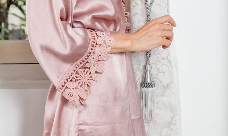 May include: A pale pink satin robe featuring floral lace detailing on the sleeves. The robe is crafted from a smooth, lustrous material, giving it a delicate and feminine look. The lace trim provides an elegant touch to the garment.