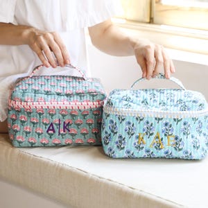 May include: Two quilted cosmetic bags with floral patterns and initial embroidery. One bag is teal with pink flowers and the initials "AK", the other is light blue with purple flowers and the initials "AAE". Both have handles.