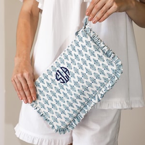 May include: A patterned clutch bag with a monogram, held by a person wearing white pyjamas. The clutch has a repeating blue floral design on a white background, with a ruffled edge and a handle.