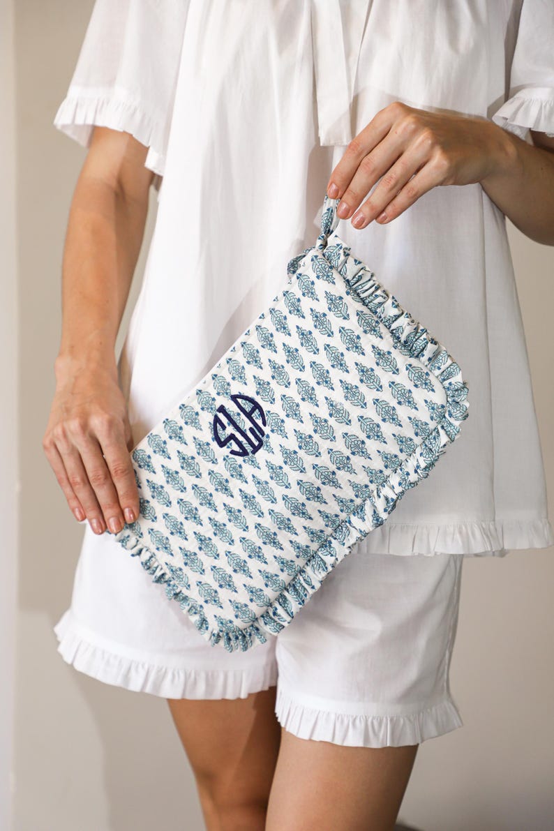 May include: A patterned clutch bag with a monogram, held by a person wearing white pyjamas. The clutch has a repeating blue floral design on a white background, with a ruffled edge and a handle.
