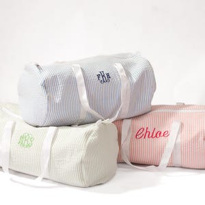 Puede incluir: Tres bolsas de lona a rayas con correas blancas. Las bolsas est&aacute;n personalizadas con los nombres "F.R.H." en azul marino, "Chloe" en rosa y un monograma verde.